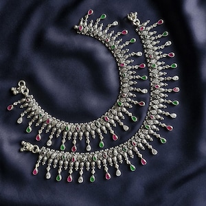 Peut inclure: Une paire de bracelets de cheville argentés présentant un design incurvé avec un travail du métal complexe. Les bracelets sont ornés de nombreuses pierres en forme de goutte pendantes aux couleurs alternées rouge, vert et blanc.