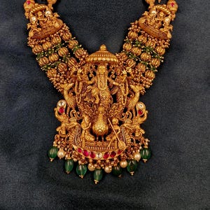 May include: A gold-toned necklace featuring a large central pendant depicting a deity figure, surrounded by intricate metalwork, small red and green stones, and dangling green beads. The chain consists of spherical gold beads and smaller deity motifs.