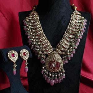 May include: A gold-toned necklace and matching earring set featuring intricate metalwork, inlaid red stones, and dangling pink teardrop beads. The necklace has a large central pendant with a stylized bird motif and small green bead accents.
