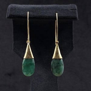 Peut inclure: Une paire de boucles d'oreilles pendantes dorées avec des pierres précieuses vertes émeraude en forme de goutte. Les boucles d'oreilles sont présentées sur un présentoir à bijoux en velours noir, mettant en valeur leur design élégant et leur couleur vibrante.