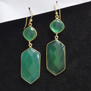 May include: A pair of green gemstone earrings with gold-coloured frames. Each earring features a square-shaped top stone and a larger, faceted, emerald-cut bottom stone. The earrings hang from gold-coloured ear wires.