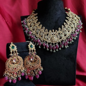 May include: A gold-toned necklace and matching pair of earrings featuring intricate metalwork, embedded clear stones, and dangling oval-shaped pink beads. The jewelry set is accented with small green beads along the lower edges and green stone inlays.