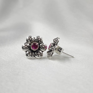 Può includere: Un paio di orecchini a bottone argentati. Ogni orecchino presenta un design floreale con una gemma rossa rotonda centrale. I petali del fiore sono formati da piccole sfere strutturate. Gli orecchini sono su sfondo bianco.