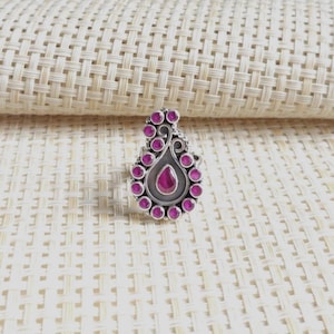 Può includere: Un anello in argento con un elemento centrale a forma di goccia, caratterizzato da una pietra rosa centrale circondata da un alone di pietre rosa rotonde più piccole. Il metallo presenta una finitura ossidata che mette in risalto i dettagli.
