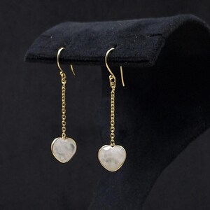 May include: A pair of gold-coloured drop earrings. Each earring has a heart-shaped, pale stone pendant hanging from a fine chain. The earrings are shown against a dark backdrop.