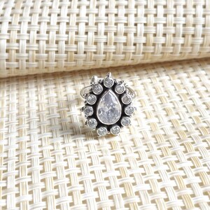 Può includere: Un anello color argento con una pietra centrale trasparente a forma di pera, circondata da un alone di dodici pietre rotonde trasparenti più piccole. La montatura in metallo presenta una finitura scura e ossidata negli spazi tra le pietre.