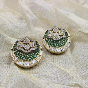 May include: A pair of gold-toned stud earrings. Each earring features a central cluster of small green stones, surrounded by a border of clear, square-cut stones. The top of each earring has a design with clear, teardrop-shaped stones.