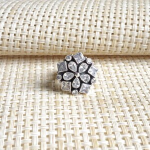 Può includere: Un anello color argento con un gruppo di pietre trasparenti sfaccettate disposte a motivo floreale. Il design presenta una pietra centrale a forma di pera circondata da pietre più piccole rotonde, quadrate e a goccia su una montatura metallica.