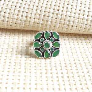 Può includere: Un anello in argento con una gemma verde rotonda centrale circondata da quattro pietre verdi a forma di marquise e quattro pietre verdi rotonde più piccole disposte a motivo floreale. L'incastonatura in metallo ha una finitura ossidata scura.