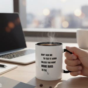 May include: White coffee mug with a black interior and handle, featuring the text "DON'T ASK ME TO TEST IT AGAIN UNLESS YOU WANT MORE BUGS TO FIX." Steam rises from the mug, suggesting a hot beverage. A laptop and notepad are in the background.