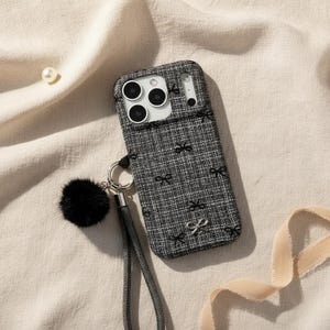 May include: A gray and white patterned phone case with black bow accents. The case has a black wrist strap and a black fluffy pom-pom charm. The phone case has a camera cutout with three lenses.