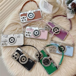 Puede incluir: Una colección de fundas para teléfono con forma de cámara en varios colores: marrón, blanco, rosa, morado, azul, verde y negro. Cada funda tiene una lente plateada y una correa. Las fundas se muestran sobre una superficie blanca y esponjosa.