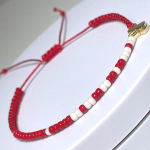 Könnte beinhalten: Armband mit roten und weißen Perlen und einem goldfarbenen Kreuzanhänger. Das Armband hat ein geflochtenes rotes Band und einen verstellbaren Schiebeknotenverschluss. Die Perlen wechseln sich in Rot und Weiß ab.