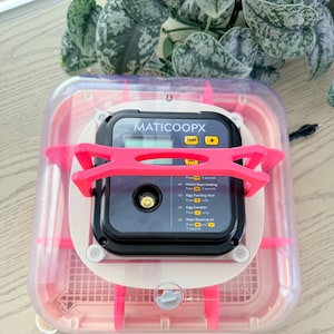 May include: A clear and pink incubator with a digital display labeled "MATICOOPX". The incubator has a pink support structure and a clear lid. The display shows temperature and other settings. A plant is visible in the background.