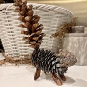 May include: A handcrafted pinecone sculpture of an animal with a tail and legs, standing in front of a white wicker basket. The sculpture is brown and gray, with a natural, rustic appearance. Decorative elements.