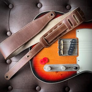 May include: A brown leather guitar strap with cream stitching is laid across a sunburst electric guitar. The strap has adjustable holes and a padded shoulder section. Two red guitar picks are visible.