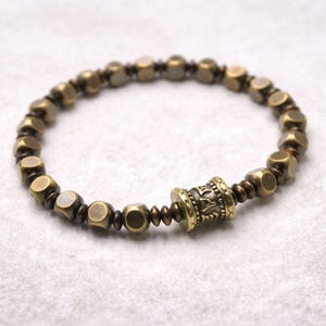 May include: A brass bracelet featuring a mix of cube and round beads. The bracelet has a central cylindrical bead with etched details. The beads are a warm, antique gold color, strung together on a dark brown cord.