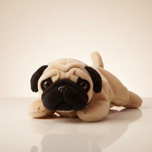 May include: A plush pug dog toy in a lying position. The toy has a beige body, black ears, and a black snout. The pug's eyes are dark and it has a small tail. The toy is on a white surface.