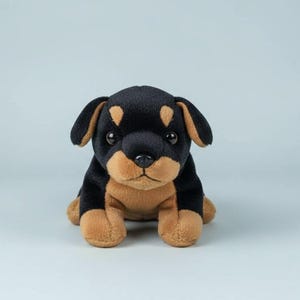 May include: A plush Rottweiler puppy toy. The stuffed animal has black fur with tan markings on its face, ears, and paws. It has a soft, cuddly appearance and is sitting upright.