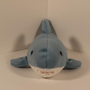 May include: A plush shark toy in shades of blue and white. The shark has a light blue body, a dorsal fin, and small side fins. Its mouth is open, revealing white teeth and a pink interior. The toy is designed to resemble a shark.