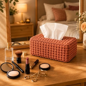 May include: A rectangular, terracotta-colored tissue box cover with a textured, woven design. White tissues are visible at the top. The box sits on a wooden vanity with makeup and a mirror. A perfume bottle and makeup brushes are also visible.