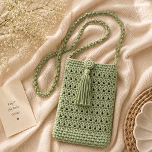 May include: A sage green crochet crossbody bag featuring a rectangular body with an openwork triangular stitch pattern, a long braided strap, and a decorative tassel attached to a circular button detail at the top center of the bag.