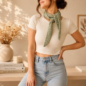 Puede incluir: Un pañuelo triangular de ganchillo en color verde salvia atado al cuello, combinado con una camiseta corta blanca de canalé con mangas cortas y bordes ondulados, y unos vaqueros azul claro con cierre clásico de botón y cremallera.