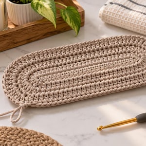 May include: An oval-shaped, beige crochet mat featuring a dense, textured stitch pattern. The mat has a small, looped cord attached to one end, providing a simple detail to the overall construction of this handmade textile piece.