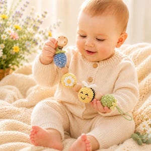 May include: A baby in a cream-colored knit outfit holds a wooden and crochet rattle. The rattle features a blue, yellow, and green design with a bee and flower motif. The baby is sitting on a cream-colored blanket.
