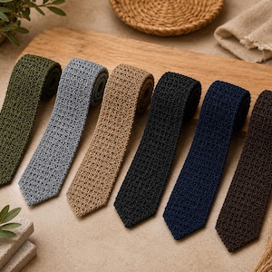Peut inclure: Une collection de six cravates en tricot déclinées en vert olive, gris clair, beige, noir, bleu marine et marron foncé. Chaque cravate présente une texture à motifs carrés uniforme et une extrémité plate et carrée, alignées côte à côte.