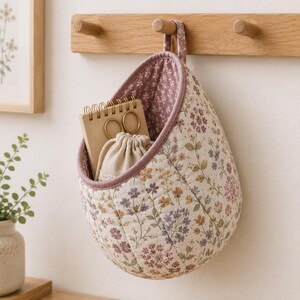 May include: A quilted fabric hanging storage basket featuring a cream background with a multi-colored floral print in shades of purple, yellow, and green. The interior is lined with a mauve floral fabric, containing a spiral notebook and a small pouch.