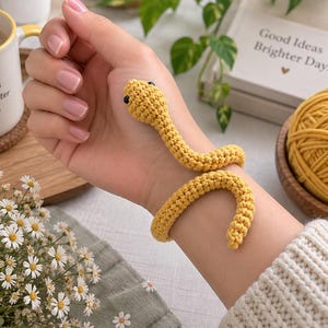 Könnte beinhalten: Ein gelbes Häkel-Schlangenarmband mit einem strukturierten, gewundenen Körper und zwei kleinen schwarzen Perlenaugen. Die Schlange ist so gestaltet, dass sie sich um das Handgelenk legt, wobei der Kopf nahe der Handfläche ruht und der Schwanz sich krümmt.