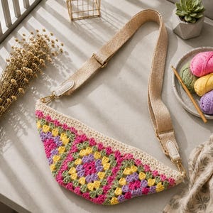 Può includere: Borsa all'uncinetto fatta a mano con bordo color crema e motivo geometrico rosa, giallo, verde e viola. Ha un cinturino beige con hardware color oro. La borsa è su una superficie grigio chiaro.