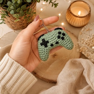 May include: A sage green crocheted video game controller keyring featuring black yarn details for the directional pad and buttons. The soft, textured yarn accessory is attached to a standard silver-toned metal split ring for easy attachment.