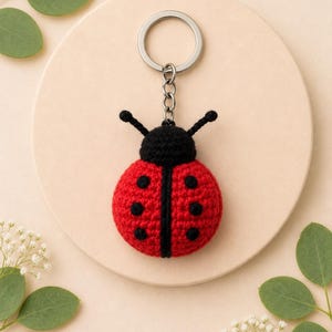 May include: A crocheted ladybird keyring featuring a round red body with black spots and a black head with two antennae. The ladybird is attached to a silver-coloured metal split ring by a short chain, creating a small, textured, handmade accessory.