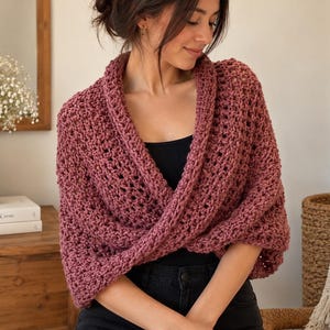 May include: A dusty rose crochet shrug featuring a textured, open-stitch pattern with a wide collar and a wrap-around front design. The garment is worn over a black top and dark pants, showcasing a thick, yarn-based construction with long sleeves.