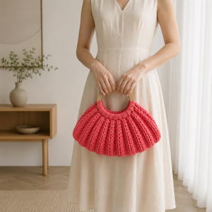 Puede incluir: Un bolso de mano color rosa coral vibrante con una textura de punto acanalado grueso y un asa circular de madera clara. El bolso tiene una silueta semicircular plisada y está confeccionado con material de hilo grueso, mostrando un patrón vertical.