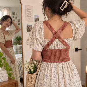 May include: A cream-colored dress with a blue floral pattern featuring puff sleeves and a square neckline, worn with a terracotta-colored crocheted corset top that has a lace-up front and wide crisscross straps on the back.