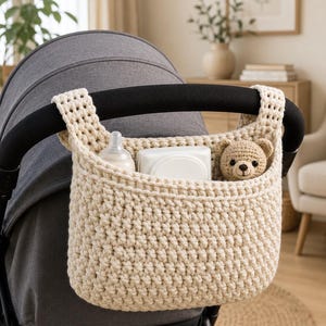 May include: A cream-colored crochet hanging basket attached to a stroller handle. The basket contains a baby bottle, a white rectangular container, and a small crocheted teddy bear with brown yarn and black eyes, providing storage for baby items.