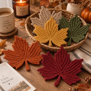 Puede incluir: Un conjunto de cinco posavasos de ganchillo con forma de hoja de arce en colores otoñales: beige, amarillo, verde oscuro, naranja y rojo intenso. Tres hojas están dentro de una cesta de mimbre redonda y dos descansan sobre una superficie de madera.