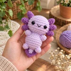 Könnte beinhalten: Ein kleines, handgefertigtes Häkel-Axolotl-Stofftier mit hellviolettem Körper, dunkelvioletten äußeren Kiemen und zwei schwarzen Kunststoffperlen als Augen. Das Wesen hat einen runden Kopf, einen prallen Rumpf, kleine Gliedmaßen und einen flachen Schwanz.