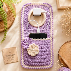 以下が含まれることがあります： ラベンダー色のクロッシェ編みスマホホルダー。上部にはクリーム色のリング、前面には2つのクロッシェ編みの花が飾られています。ホルダーのポケットには黒いスマートフォンが入っており、白い充電ケーブルと白い電源アダプターに接続されています。