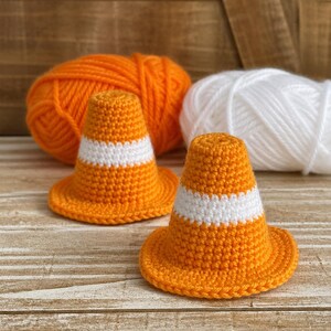May include: Two handmade crochet traffic cones in bright orange with a white stripe. The cones are on a wooden surface, with a ball of orange yarn and a ball of white yarn in the background. The cones are a fun, whimsical craft.