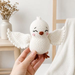 Op de afbeelding: Een handgemaakte witte gehaakte vogel met uitgestrekte vleugels. De vogel heeft een bruine snavel en poten, met roze wangen en gesloten ogen. De vleugels hebben een geschulpte rand en de vogel lijkt te glimlachen.