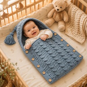 May include: A blue crocheted baby sleep sack with a hood, a decorative tassel, and wooden buttons along the sides. The sleep sack features a textured stitch pattern and is designed to wrap around a baby, providing a cozy and secure covering.