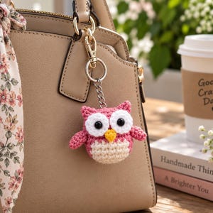 May include: A pink and cream crocheted owl keychain with large white eyes, black pupils, and a yellow beak, attached to a gold-toned metal clasp. The owl features a textured yarn body and is hanging from a beige handbag adorned with a floral scarf.