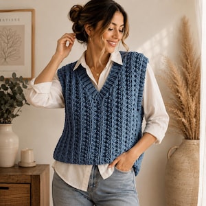 May include: A blue knitted sweater vest featuring a V-neckline and a vertical ribbed texture, worn over a crisp white long-sleeved button-down shirt and light blue denim jeans. The vest has a thick, chunky knit construction with a sleeveless design.