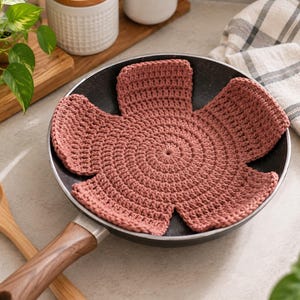 Puede incluir: Un tapete de ganchillo de color rosa polvoriento, circular y con seis extensiones en forma de pétalo, colocado dentro de una sartén negra. El tapete presenta un patrón de puntos densos y texturizados que irradian desde el centro.