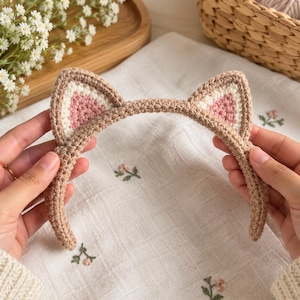 May include: A crochet headband featuring two pointed cat ears. The headband is made of light brown yarn, while the inner sections of the ears are crafted with white and soft pink yarn, creating a distinct contrast in colour and texture.