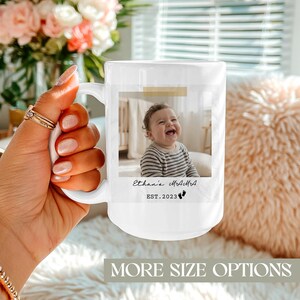 Puede incluir: Taza de cerámica blanca con una foto de un bebé, el texto "Ethan's MAMA" y "EST. 2023". La taza es sostenida por una persona. El fondo incluye un ramo de flores y un cojín esponjoso. La taza tiene un asa.